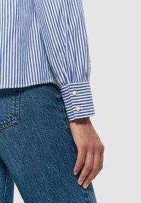 Camicia a righe blu e bianche con polsini abbottonati, abbinata a un paio di jeans; la camicia presenta maniche lunghe e un design con colletto classico.