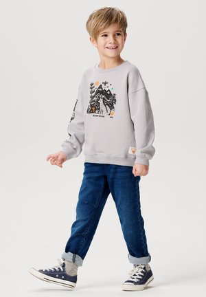 Grauer Pullover mit Grafik von Bergen und Natur, kombiniert mit dunkelblauen Jeans und weißen Turnschuhen. Hochgekrempelte Säume an den Jeans, subtile Textur.