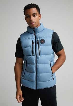 Uomo che indossa un gilet imbottito azzurro chiaro con tasche con cerniera frontali e sul petto sopra una maglietta nera a maniche corte su sfondo grigio uniforme.