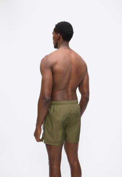 Μυώδης άνδρας που στέκεται με την πλάτη, φοράει ελαφρώς υφασμένα χακί σορτς με ελαστική μέση, πάνω σε απλό φωτεινό φόντο.