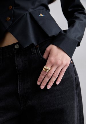 Black button-up shirt with a pleated design, featuring a small colorful embroidered patch. Hand rests on dark denim jeans, showcasing gold rings.