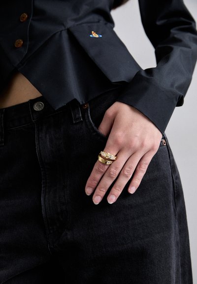 Schwarze Hemd mit Knöpfen und einem gefalteten Design, versehen mit einem kleinen bunten gestickten Aufnäher. Die Hand ruht auf einer dunklen Jeanshose und zeigt goldene Ringe.