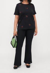 Svart t-shirt med röda rosdekorationer, ihopparat med svarta utsvängda byxor. Håller en grön och svart pärlhandväska. Platta svarta skor.