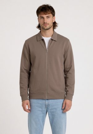 Mężczyzna w taupe zapinanej na zamek kurtce, białej koszuli i jasnoniebieskich dżinsach, stojący na tle jednolitego jasnego tła.