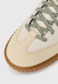 Beige sneakers med semskede og lærpaneler, gummigummisåle, og grønne og hvide mønstrede flade snørebånd bundet i krydsmønster.