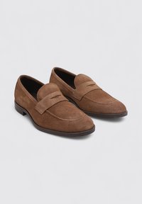Mocassins en daim marron avec un talon plat, présentant des détails cousus sur le bout et une bande à l'avant.