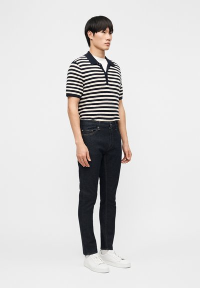 Mladý muž stojící, oblečený v černobílém pruhovaném krátkém polo tričku, tmavých džínách, bílých ponožkách a bílých teniskách, na jednobarevném pozadí.