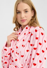 Albornoz de forro polar en rosa claro con estampados de corazones rojos, que presenta una textura suave, diseño cruzado y un cinturón para atar en la cintura.