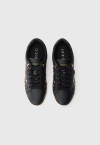 Sneakers nere Guess con occhielli dorati e logo, lacci neri e dettagli a stampa leopardata sul tallone, viste dall'alto su sfondo bianco.