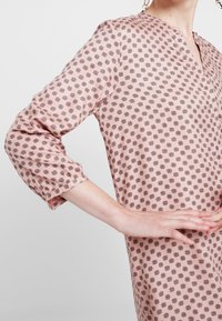 Růžové šaty s vzorem a tříčtvrtečními rukávy, které mají opakující se tmavý květinový motiv, lehký materiál a výstřih do V.
