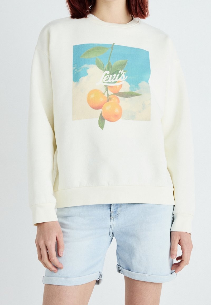 Personne portant un sweat-shirt blanc avec un motif de fruit orange et feuille et un short en denim bleu clair retroussé.