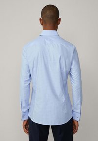 Camicia a quadratini azzurri chiari con maniche lunghe, polsini con bottone e colletto classico. Presenta un orlo curvo e una texture di tessuto liscia.