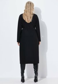 Zwarte lange jurk met lange mouwen en een taille met riem, gemaakt van een gladde stof. Gemonteerd met knie-hoge zwarte laarzen. De achteraanblik laat een ontspannen pasvorm zien.