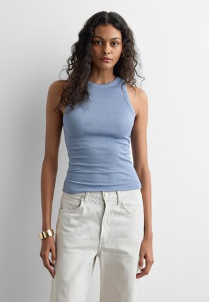 Jonge vrouw met lang krullend haar, gekleed in een lichtblauw, geribd, mouwloos topje, witte broek en een gouden armband, staand tegen een witte achtergrond.