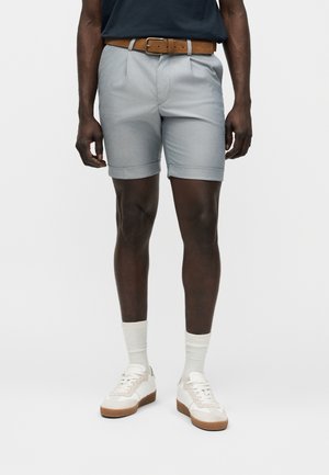 Άνδρας που φοράει ανοιχτογκρι tailored σορτς με γυρισμένα τελειώματα, καφέ δερμάτινη ζώνη, λευκές κάλτσες και λευκά αθλητικά παπούτσια, στέκεται μπροστά σε ένα μονόχρωμο φόντο.