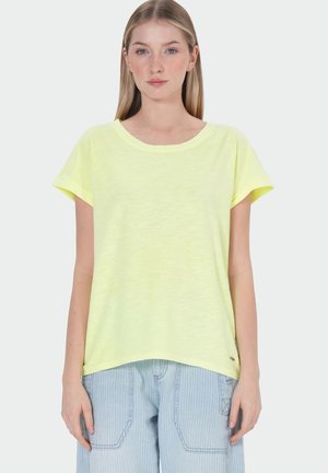 Jeune femme portant un haut jaune pâle à manches courtes et un pantalon rayé bleu clair, debout sur un fond blanc.