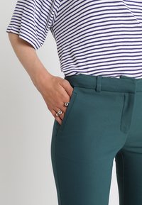 Randig vit och lila T-shirt kombinerad med turkosa tajta byxor, med sidofickor och en mjuk yta. Handen vilar på fickan.