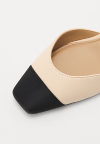 Chaussure à talon bloc beige et noir avec un dessus en cuir lisse, ouverture élastiquée et bout carré. Le talon texturé noir contraste avec le dessus.
