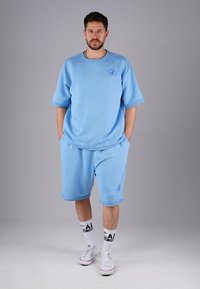 Light blue cotton tracksuit featuring a relaxed-fit T-shirt and shorts, with a round neckline and logo detail. Worn with white sneakers.