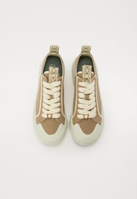 Baskets en toile marron avec semelles en caoutchouc crème, mettant en avant des lacets crème, un accent doré près de la languette et une étiquette à motif au niveau du talon.