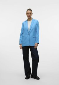 Blazer azzurro chiaro con un solo bottone, revers a punta e due tasche frontali, abbinato a pantaloni in denim scuro e scarpe nere.