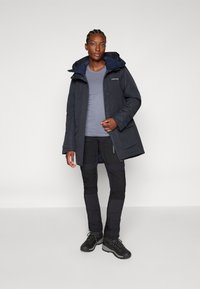 Marineblauwe waterdichte jas met capuchon, gedragen met een grijze top en zwarte broek. Voorzien van ritszakken, versterkte knieën en stevige zwarte schoenen.