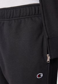 Pantalones de chándal negros con una textura suave, cinturilla elástica y un pequeño logotipo bordado en rojo y blanco. Incluye un detalle de bolsillo con cremallera.
