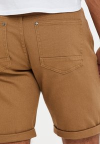 Pantalones cortos de mezclilla marrones con puños enrollados, que cuentan con dos bolsillos traseros con parche asegurados con botones de metal y una superficie texturizada.