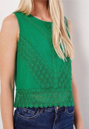 Ærmeløs grøn top med blomstrede blonde detaljer båret med blå jeans, med langt blondt hår over den ene skulder.