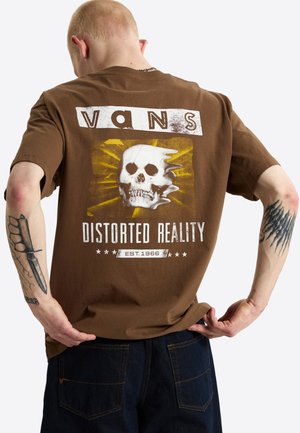 Person mit kurzen Haaren trägt ein braunes Vans-T-Shirt mit Totenkopf-Grafik und dem Schriftzug "Distorted Reality" auf dem Rücken und zeigt Tattoos auf beiden Armen.