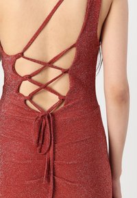 Robe rouge scintillante avec un dos échancré, présentant des bretelles croisées et un détail à nouer à la taille. Tissu lisse et ajusté avec une texture chatoyante.