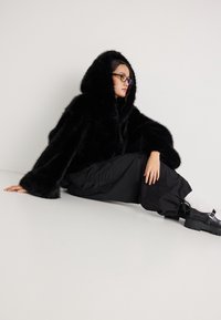 Juodas dirbtinės kailio gobtuvu megztinis su plačiomis rankovėmis, dėvimas ant juodų kelnių ir storapadžių batų. Lygus paviršius su blizgiu apdaila.