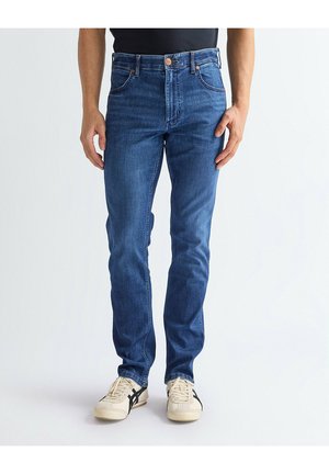 Mężczyzna w dopasowanych niebieskich dżinsach i białych tenisówkach z czarnymi paskami, stojący na tle jednolitego, jasnego tła.