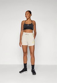 Mulher de pé e a sorrir, vestindo um top desportivo preto, calções bege com riscas amarelas, meias pretas e sapatilhas pretas.