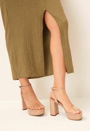 Sandales à plateforme couleur nude avec une lanière transparente, un talon bloc et une bride autour de la cheville. La semelle est lisse et brillante, tandis que le talon est de forme carrée.