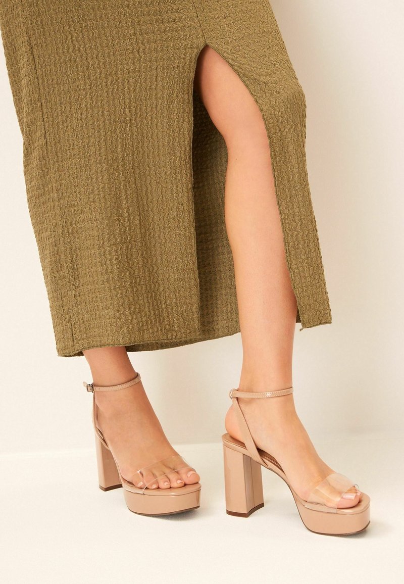 Sandales à plateforme couleur nude avec une lanière transparente, un talon bloc et une bride autour de la cheville. La semelle est lisse et brillante, tandis que le talon est de forme carrée.