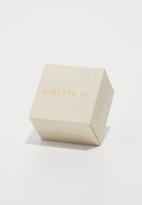 Kvadratna kutija za nakit od teksturiranog bež materijala sa zlatnim utisnutim logotipom "GINETTE NY" na poklopcu.