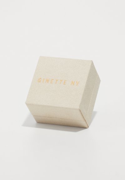 Neliömäinen korurasi, joka on valmistettu teksturoidusta beigeistä materiaalista, jossa on kultapainettu "GINETTE NY" -logo kannessa.