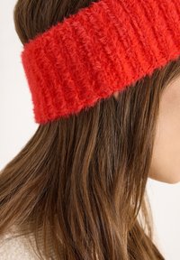 Czerwona dzianinowa opaska na głowę o puszystej fakturze i prążkowanym wzorze, noszona nad długimi, brązowymi włosami. Widok z bliska podkreślający kolor i materiał.