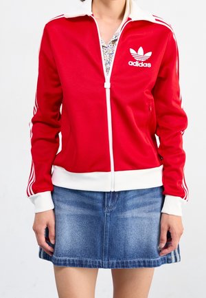 Osoba nosząca czerwoną kurtkę Adidas na zamek błyskawiczny z białymi paskami i niebieską dżinsową spódnicę, stojąca na białym tle.