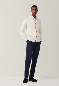 Cardigan lungo di colore crema chiaro con bottoni neri, abbinato a pantaloni blu navy. Scarponi marroni completano l'outfit. Tessuto liscio in tutto il capo.