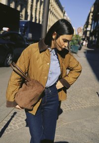 Femme aux longs cheveux noirs portant une veste matelassée marron, une chemise en jean bleue et un jean, tenant un sac marron dans une rue ensoleillée de la ville.