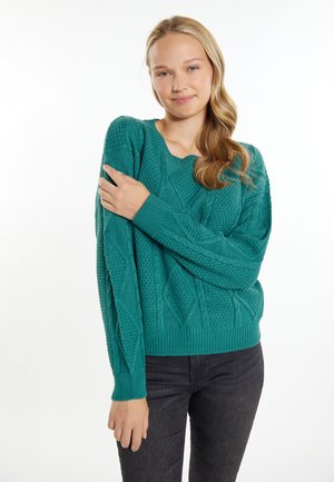 Tropfenaquafarbenes, kabelgestricktes Sweatshirt mit Rundhalsausschnitt, strukturiertem Muster und gerippten Bündchen und Saum. Das Modell kombiniert es mit dunklen Jeans.