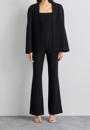 Femme debout en intérieur portant une combinaison noire évasée et un blazer assorti ouvert, sandales à talons couleur chair, pose neutre devant un mur uni.