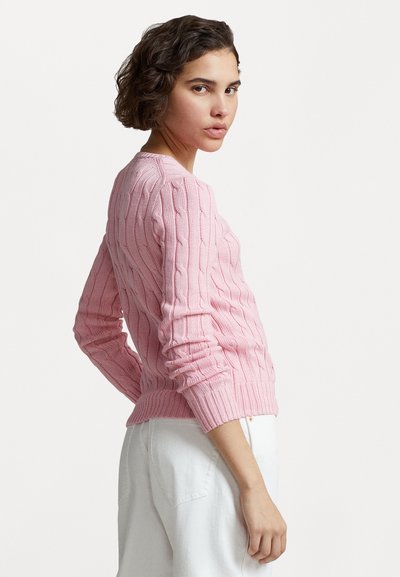 Pull en tricot rose avec un col rond et des poignets côtelés, présentant un motif texturé et une coupe ajustée, porté avec un pantalon blanc.