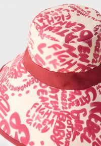 Sombrero tipo bucket de lona con texto rojo en estilo manuscrito y una banda roja sólida alrededor de la corona, con frases como "Lucha como una chica."