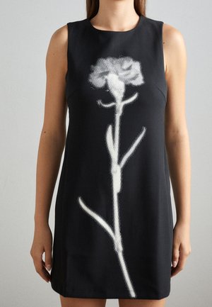 Frau trägt ein ärmelloses schwarzes Kleid mit großem weißem pixelartigen Blumenmotiv vorne vor einfarbigem Hintergrund.