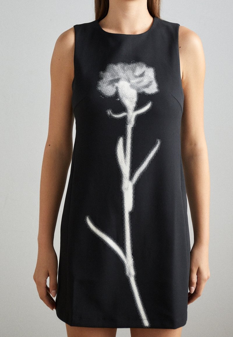 Femme portant une robe noire sans manches avec un grand motif fleur pixellisé blanc sur le devant, sur un fond uni.