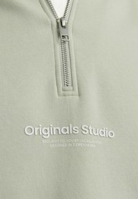 Light green quarter-zip sweatshirt with a smooth texture. Features a silver zipper and white printed branding on the front.