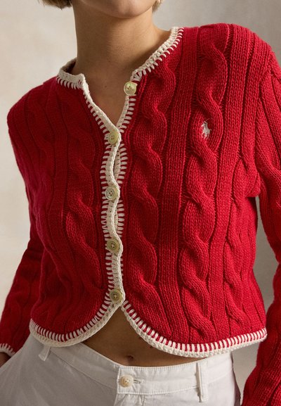 Czerwony kardigan z dzianiny warkoczowej z białym obszyciem i guzikami, noszony na białych wysokich spodniach, odsłaniający talię i szyję.